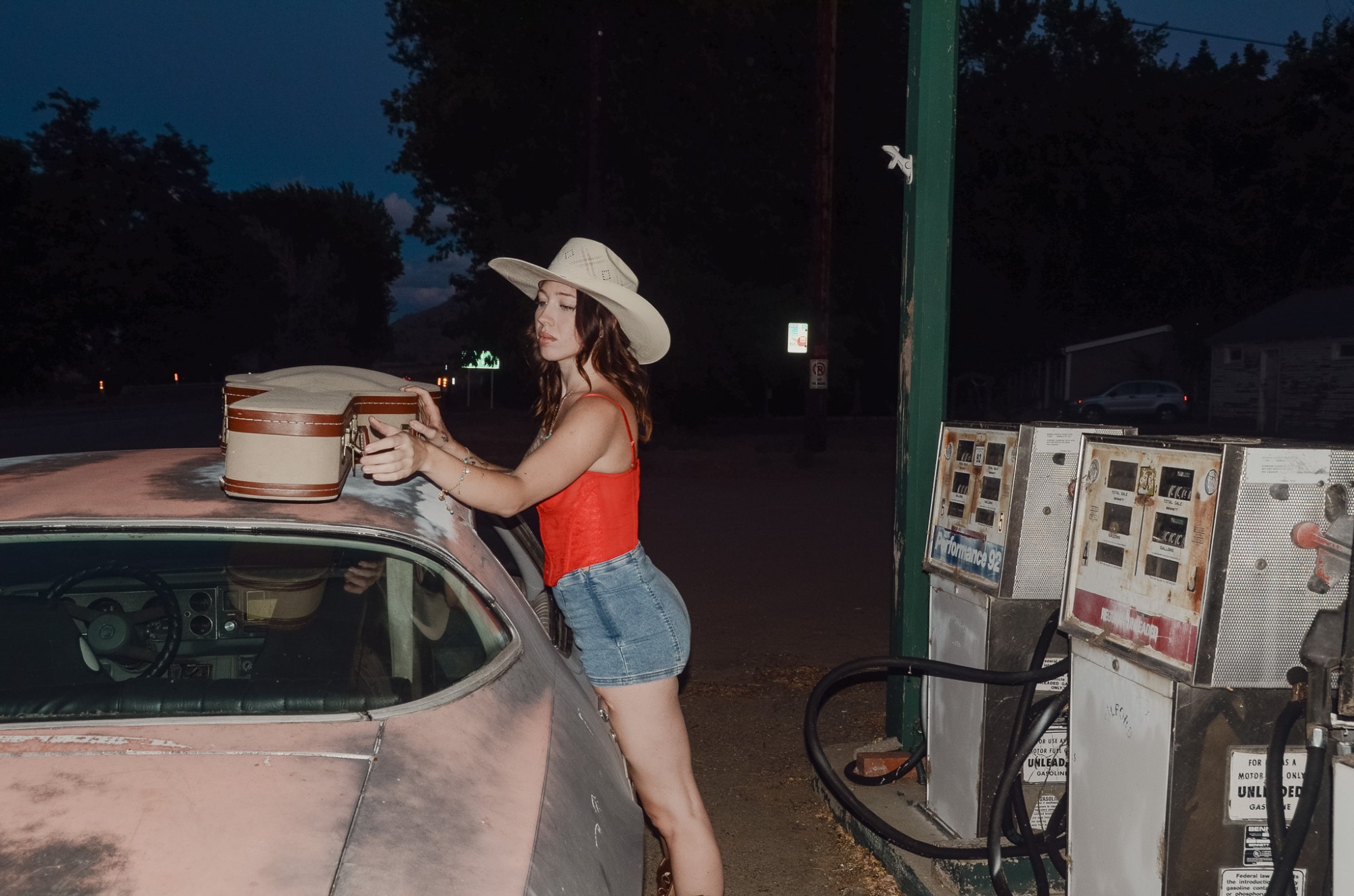 Olivia loading a vintage guitar case onto the roof of an old car beside rusted gas pumps at night