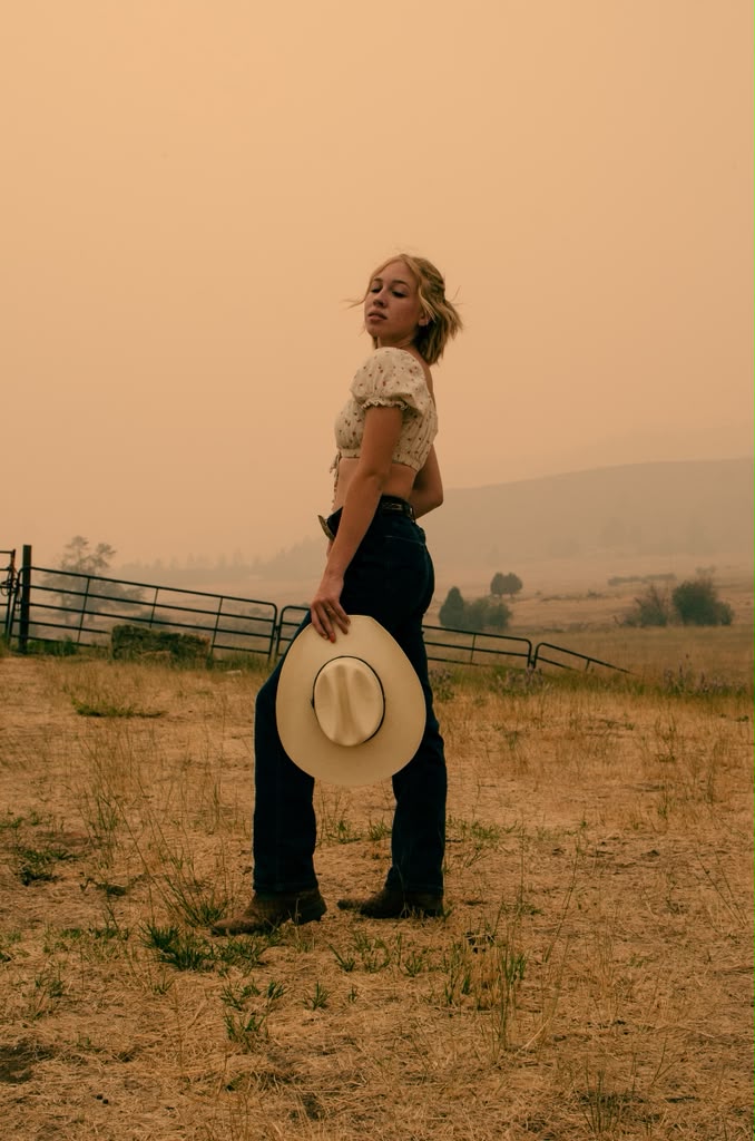 Olivia in a hazy golden field, looking back over her shoulder with a cowboy hat in hand