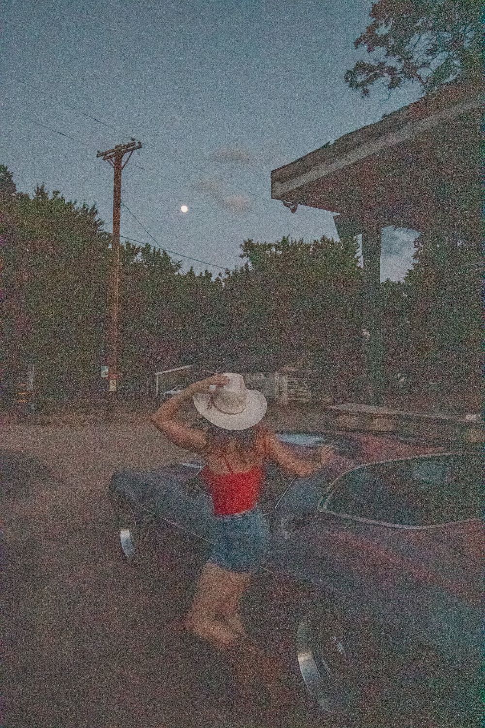 Olivia from behind, holding her hat, leaning on an old car under the moon
